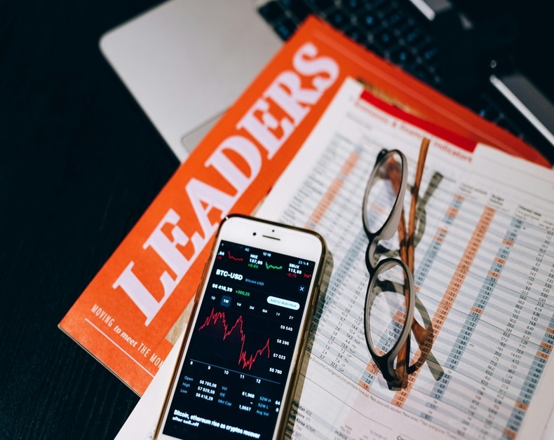 Stock market app open on a phone beside a financial magazine and reading glasses on a chart-filled document near a laptop.