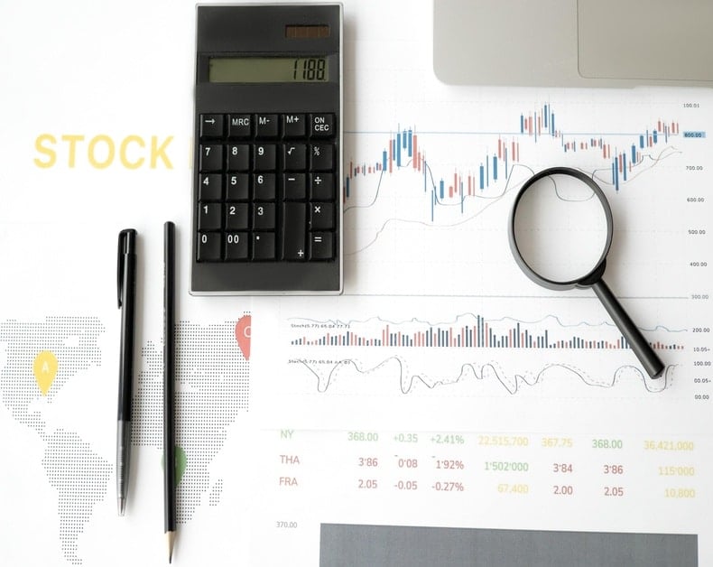Stock market analysis scene with a calculator, magnifying glass over a financial chart, and two pens on the page showing stock data and trends.
