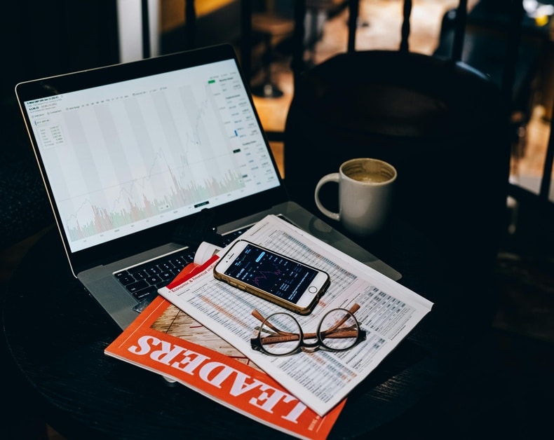 Laptop with stock charts on screen, smartphone showing a trading app, glasses, and a coffee mug on a desk with papers and a magazine nearby.