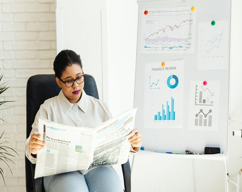 Woman with glasses reads a newspaper while seated; a whiteboard with charts and a finance section is visible behind her.