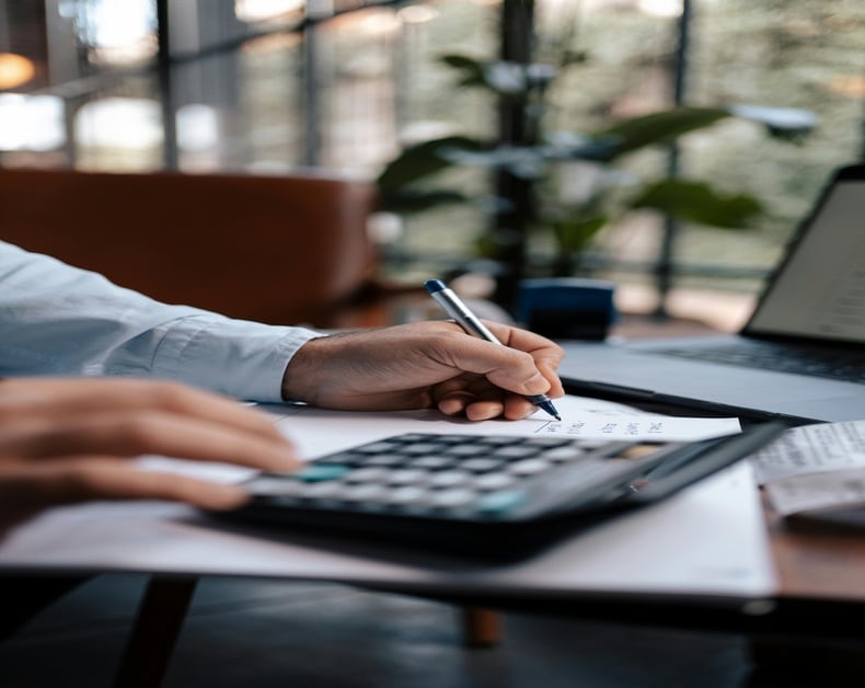 Person writes on a paper with pen at a desk, calculator and laptop nearby in a bright office.