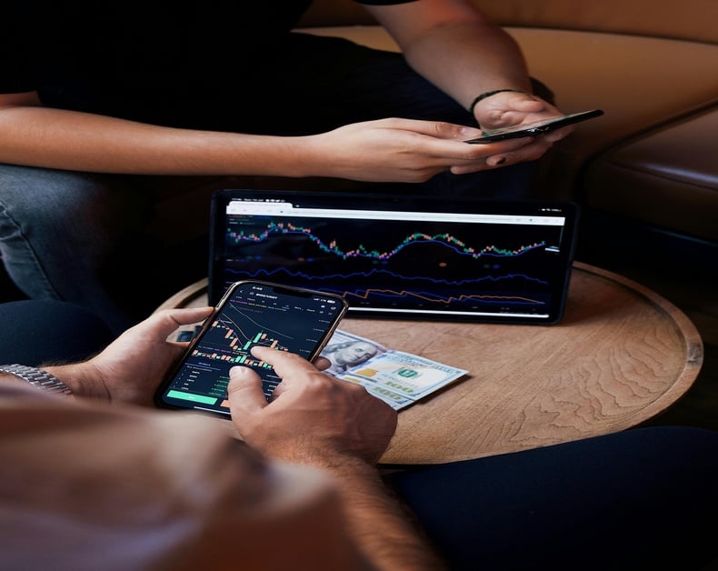 Two people check stock market charts on a tablet and a smartphone, with cash on the table.