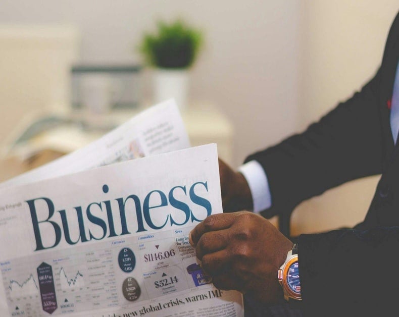 Man in a dark suit reads a large newspaper titled 'Business' at a desk, with a visible wristwatch on his left wrist.