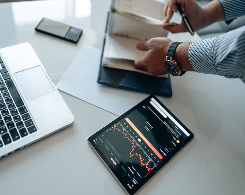 Hands open a binder on a desk, with a tablet displaying a red line graph beside a laptop.