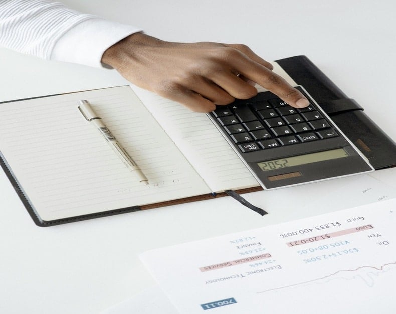 Hand using a calculator beside an open notebook with a pen and financial documents on a white desk to count costs.
