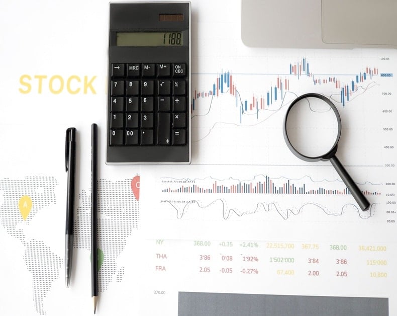 Stock analysis setup: calculator, magnifying glass, and pens over stock charts and a laptop edge.