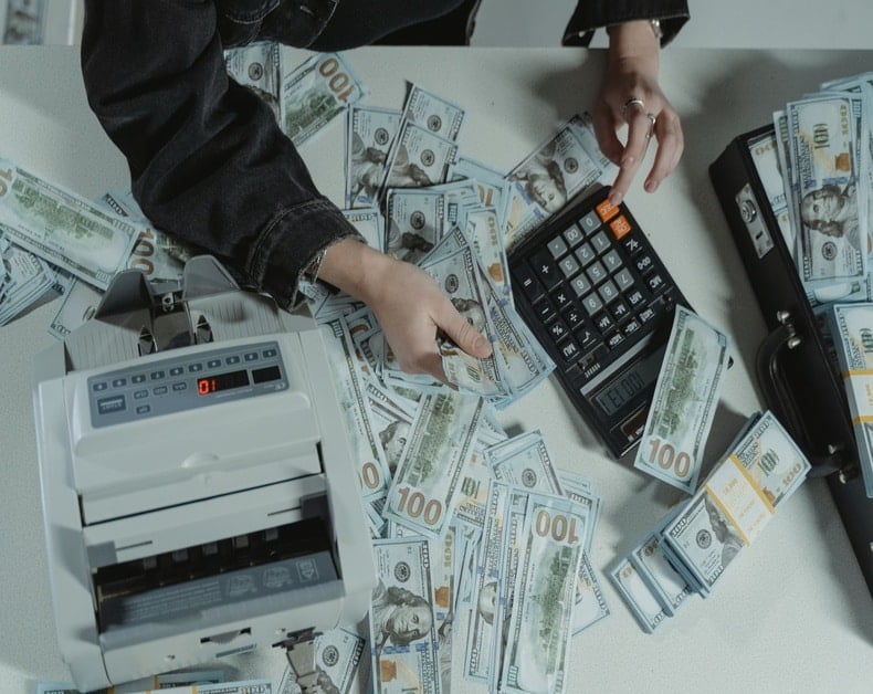 Person counts bundles of $100 bills on a desk with a money-counting machine and calculator.