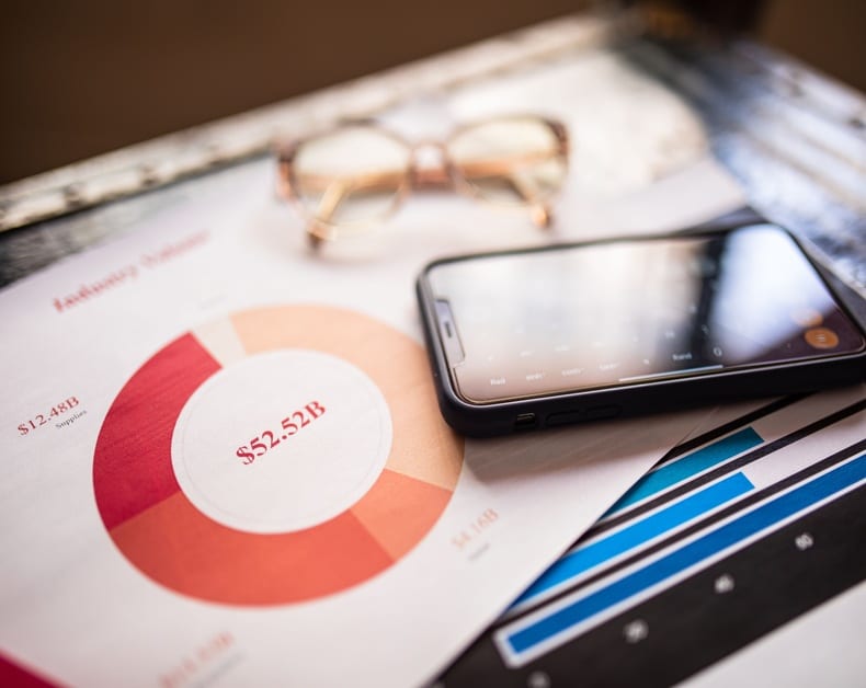 Close-up of a business report with a donut chart labeled $52.3B, a smartphone, and glasses on a desk.